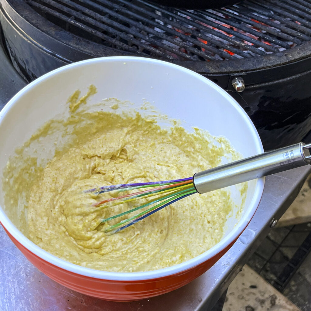 A close up of cornbread batter, in a bowl, next to a grill.