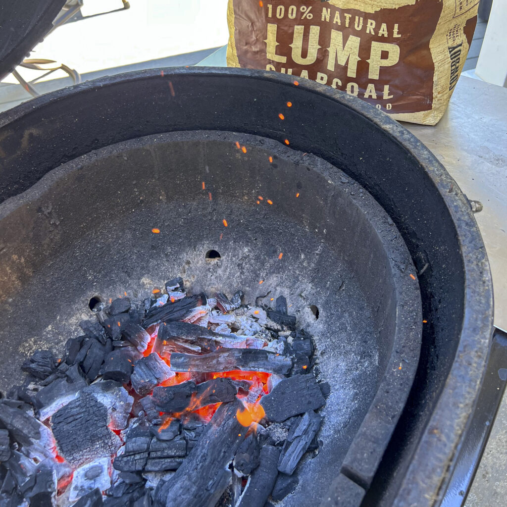 A bag that says 100% Natural Lump Charcoal is next to the grill with hot coals in it.