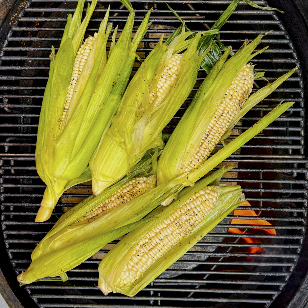 Five ears of corn, in husks, are on grill grates before roasting.
