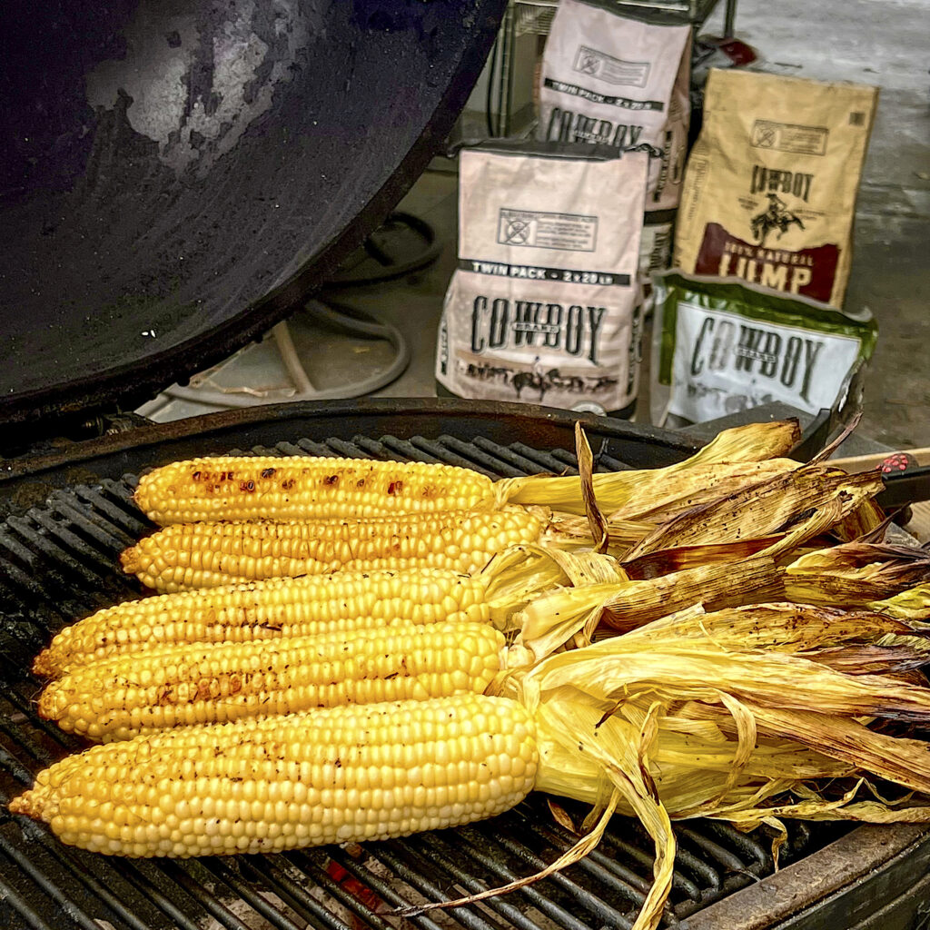 A selection of Cowboy Charcoal is in the backdrop. Five Corn on the Cob, with husks peeled back, are in the front.