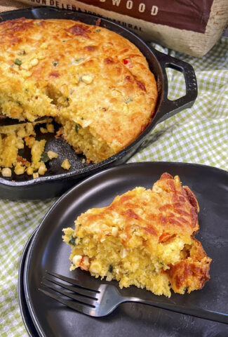 A high angle shot of a slice of cornbread on a black plate with a black fork. There is a cornbread skillet in the background on a green checkered cloth.