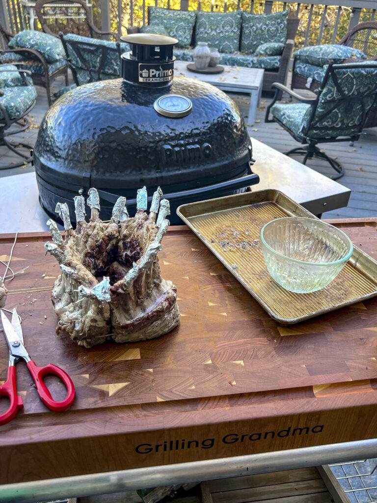 Scissors and crown of lamb are on a cutting board in front of a grill. 