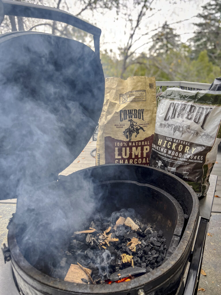 Cowboy Lump Charcoal and Cowboy Hickory Smoking Wood Chunks are near a Primo Grill that has just been loaded with lump charcoal and wood chunks. 