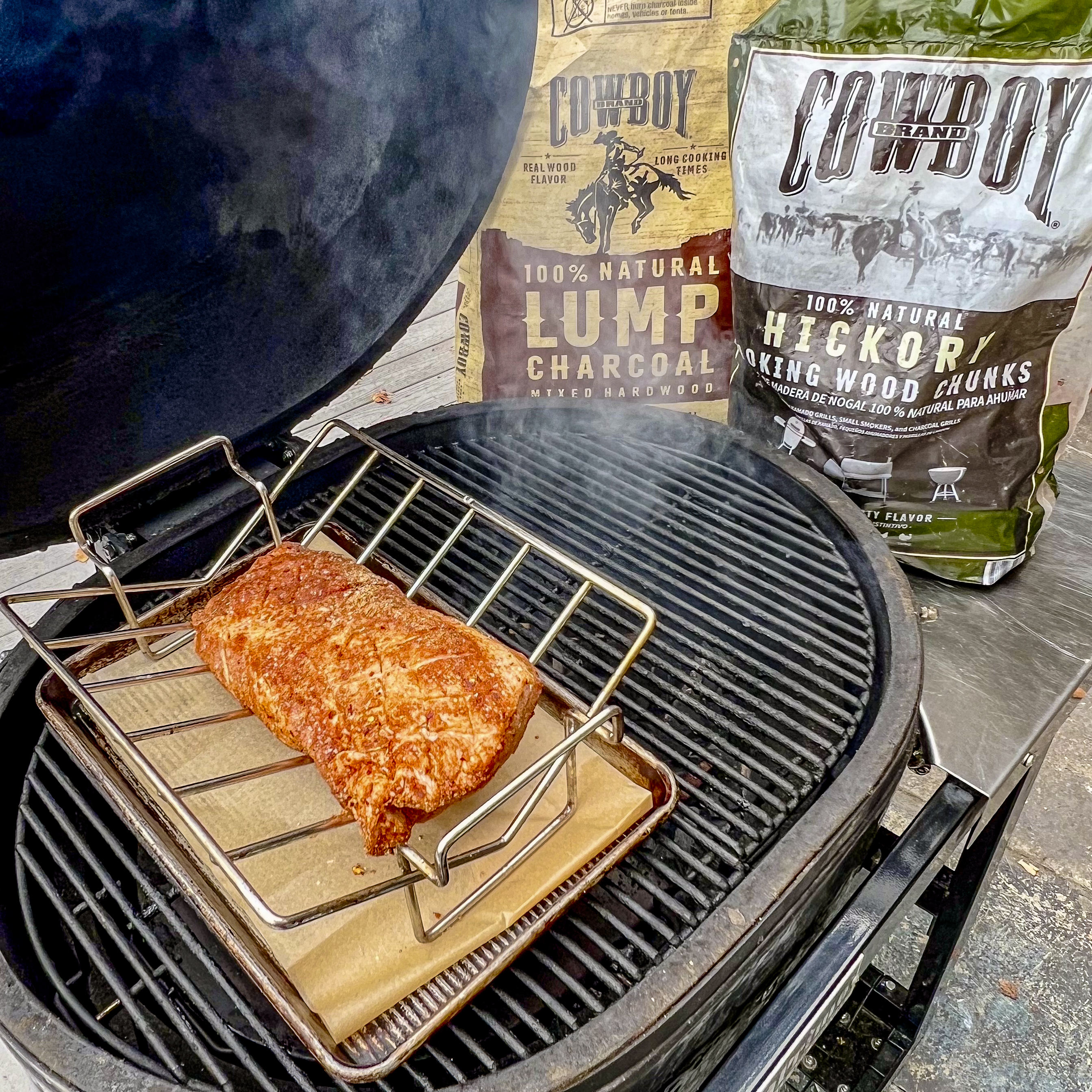 A seasoned pork loin is in a V-rack on the grill ready to smoke. A bag of Cowboy Lump Charcoal and a bag of Hickory Smoking Wood Chunks  is in the back. 