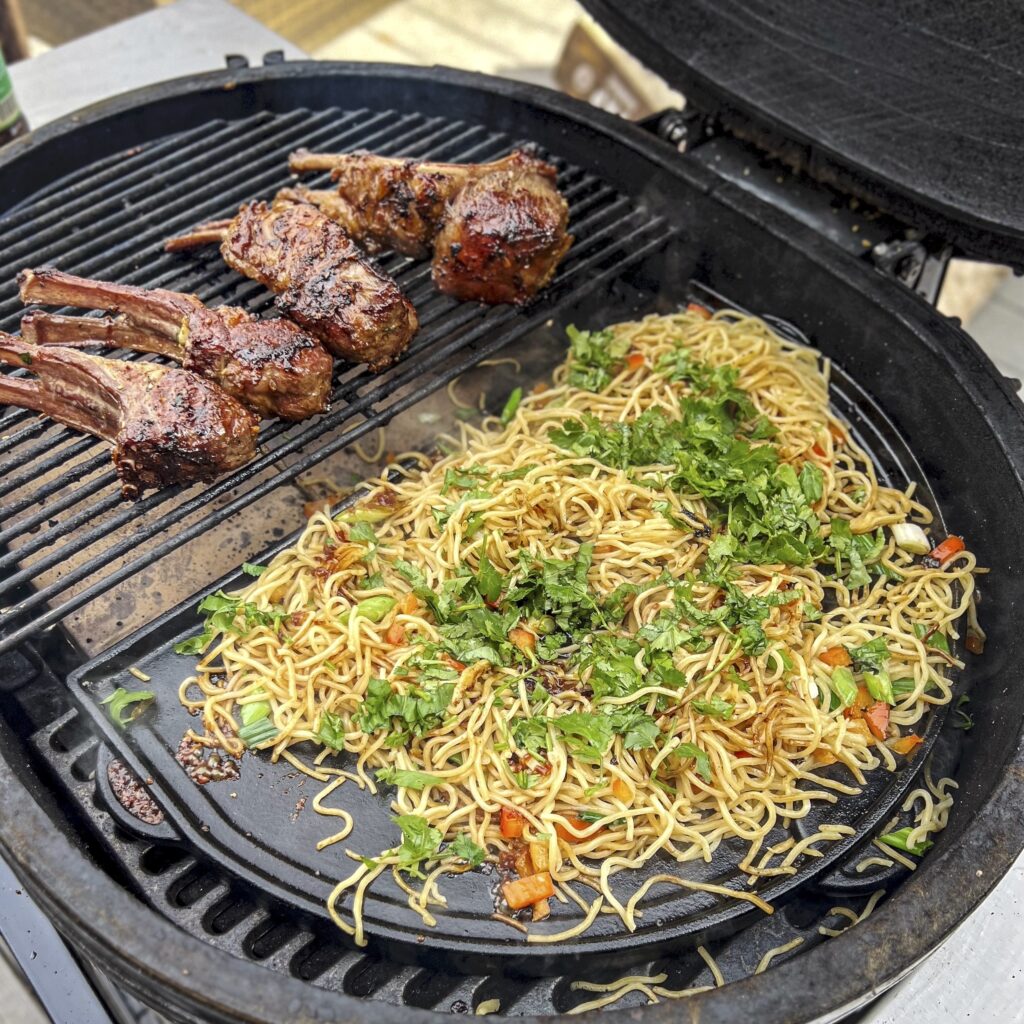 A griddle with ramen being stir-fried while lamb chops are in the background. 